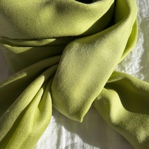May include: A close-up of a lime green scarf, draped and folded, with visible texture. The scarf is made of a soft fabric and is set against a white background. The scarf's color is vibrant and eye-catching.