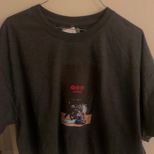 Könnte beinhalten: Dunkelgraues T-Shirt mit einem rechteckigen Aufdruck auf der Vorderseite. Der Aufdruck zeigt den Text "ose" in Rot, das Wort "ARIANAS" in Weiß und ein Foto einer Gruppe von Personen. Das T-Shirt hängt an einem Metallbügel.