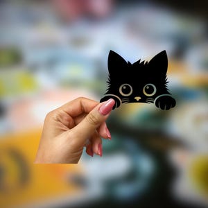 May include: A black cat silhouette sticker with large eyes and a cute expression. The sticker is being held by a hand.