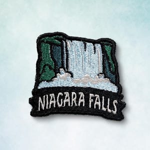 May include: Embroidered patch featuring Niagara Falls. The patch depicts a waterfall with white water cascading over a cliff, framed by green and black details. The words "Niagara Falls" are embroidered in white on a black banner.