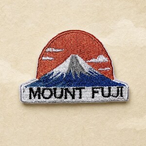 Può includere: Toppa ricamata raffigurante il Monte Fuji con un sole rosso, nuvole bianche e una base di montagna blu. Il testo "MOUNT FUJI" è ricamato in bianco su uno stendardo rettangolare. La toppa è su sfondo beige.