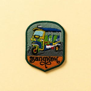 Könnte beinhalten: Gestickter Aufnäher mit einem farbenfrohen Tuk-Tuk, einem dreirädrigen Taxi, mit dem Wort "Bangkok" in einer dekorativen Schriftart. Der Aufnäher hat einen grünen Rand und einen orangefarbenen Sockel auf cremefarbenem Hintergrund.