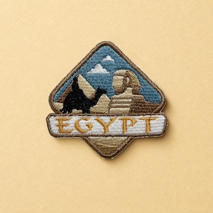 May include: A diamond-shaped embroidered patch featuring a blue sky with white clouds, a black silhouette of a person riding a camel, the Great Sphinx, and pyramids. A white horizontal banner at the bottom displays the gold text EGYPT.