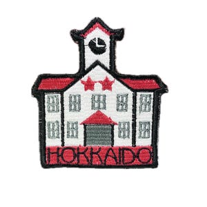 Op de afbeelding: Een geborduurde patch met een rood en wit schoolgebouw met een klokkentoren en het woord "HOKKAIDO" eronder.