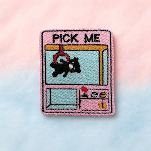 May include: Embroidered patch with a pink claw machine design and the text "PICK ME". The machine has a blue interior with a black bear held by the claw. A red button and coin slot are visible.