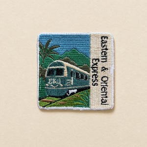 Op de afbeelding: Een vierkante geborduurde patch met een groenblauwe trein met het opschrift E&O aan de voorzijde, rijdend door een groen landschap met palmbomen en bergen. Rechts bevindt zich een crèmekleurig verticaal paneel met de tekst Eastern & Oriental Express.