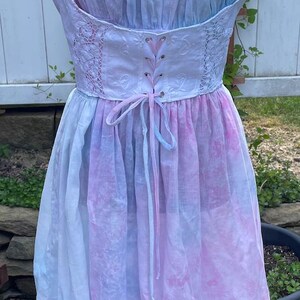 May include: A knee-length dress featuring a pastel tie-dye pattern in shades of pink and blue. The bodice includes a white lace-up corset section with pink ribbon, a gathered scoop neckline, and a hemline detailed with a white eyelet trim.