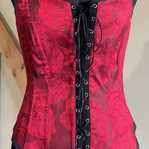 May include: A red corset with black lace-up detailing and a floral pattern. The corset is on a black mannequin.