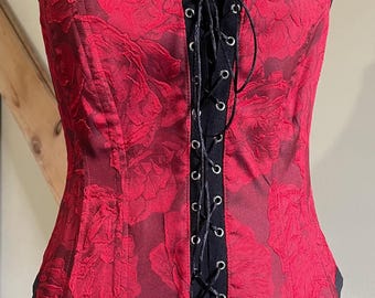 May include: A red corset with black lace-up detailing and a floral pattern. The corset is on a black mannequin.