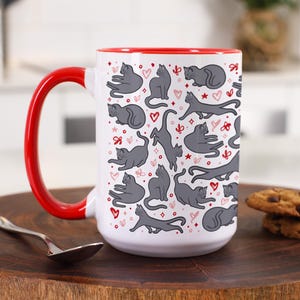 May include: A white ceramic mug with a bright red handle and interior rim. The exterior features a repeating pattern of grey cats in various poses, interspersed with small red and pink hearts, stars, and abstract floral shapes on a white background.
