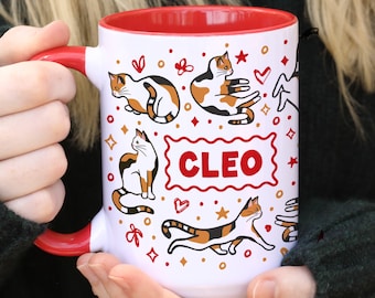 Könnte beinhalten: Ein weißer Keramikbecher mit leuchtend rotem Innenraum und Henkel, verziert mit einem wiederkehrenden Muster aus Glückskatzen in verschiedenen Posen, kleinen roten Herzen und goldenen Sternen. Der Name CLEO steht in roten Großbuchstaben.