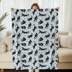 May include: A blue and white throw blanket featuring a black cat design. The cats are depicted in a variety of poses, including sitting, standing, and leaping. The blanket is soft and comfortable, ideal for curling up on the sofa.