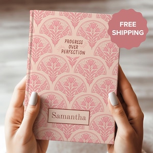 Op de afbeelding: Een roze en crèmekleurig notitieboek met een bloemenpatroon en de tekst "Progress over Perfection". Het notitieboek heeft een gepersonaliseerde naam, "Samantha", op de voorkant.