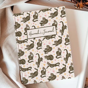 May include: A hardcover journal with a light pink background and a repeating pattern of cartoon cats in various poses. The cats are brown and gray with black stripes. The journal has the text "Hannah's Journal" on a rectangular label.