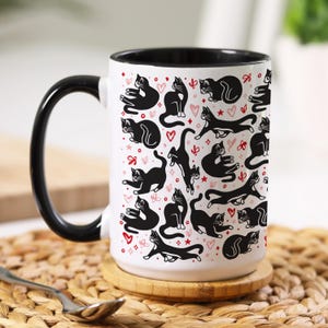 May include: White ceramic coffee mug with a black rim and handle. The mug features a repeating pattern of black cats and pink hearts.