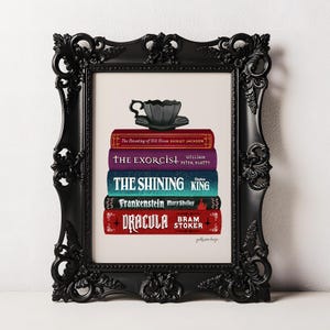 May include: A black ornate frame containing an illustration of a black teacup resting on a stack of five books titled The Haunting of Hill House, The Exorcist, The Shining, Frankenstein, and Dracula, with author names printed on each book spine.