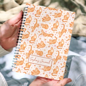 May include: A cream-coloured spiral-bound journal with a repeating pattern of orange tabby cats. The cats are in different poses, with small pink hearts and butterflies. The journal has the text "Sasha's Journal" in a rectangular box.
