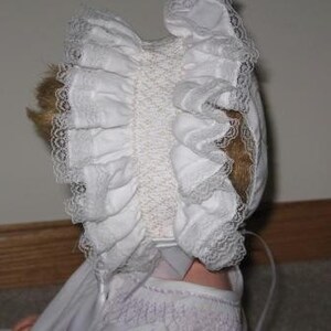 May include: A white baby bonnet with ruffled edges and lace trim. The bonnet is made of a soft fabric and has a delicate, vintage appearance. The bonnet is designed to fit a baby's head.