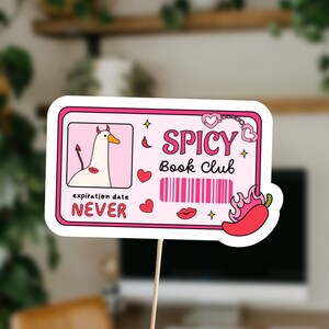 May include: A pink and white sign on a wooden stick with the words "Spicy Book Club". The sign features a cartoon goose with devil horns, hearts, a barcode, a chili pepper, and the words "expiration date NEVER".