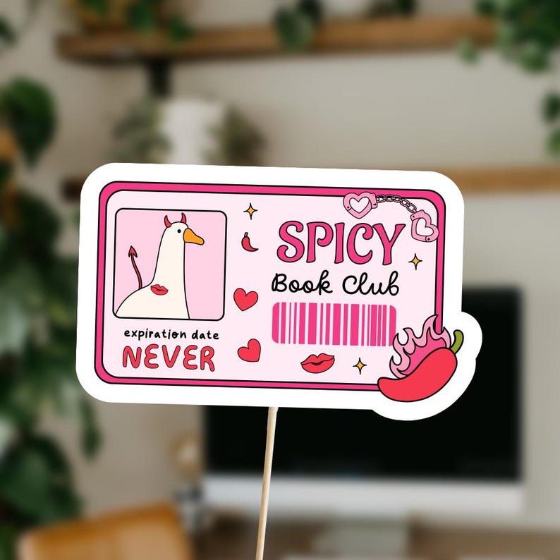 May include: A pink and white sign on a wooden stick with the words "Spicy Book Club". The sign features a cartoon goose with devil horns, hearts, a barcode, a chili pepper, and the words "expiration date NEVER".