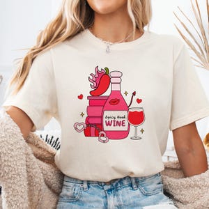 May include: Cream-colored t-shirt featuring a graphic of a wine bottle, books, and a wine glass. The bottle is pink with the text "Spicy Book Wine". Other graphics include a chili pepper, hearts, and a devil's tail.