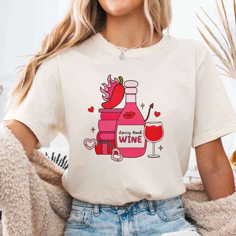 May include: Cream-colored t-shirt featuring a graphic of a wine bottle, books, and a wine glass. The bottle is pink with the text "Spicy Book Wine". Other graphics include a chili pepper, hearts, and a devil's tail.