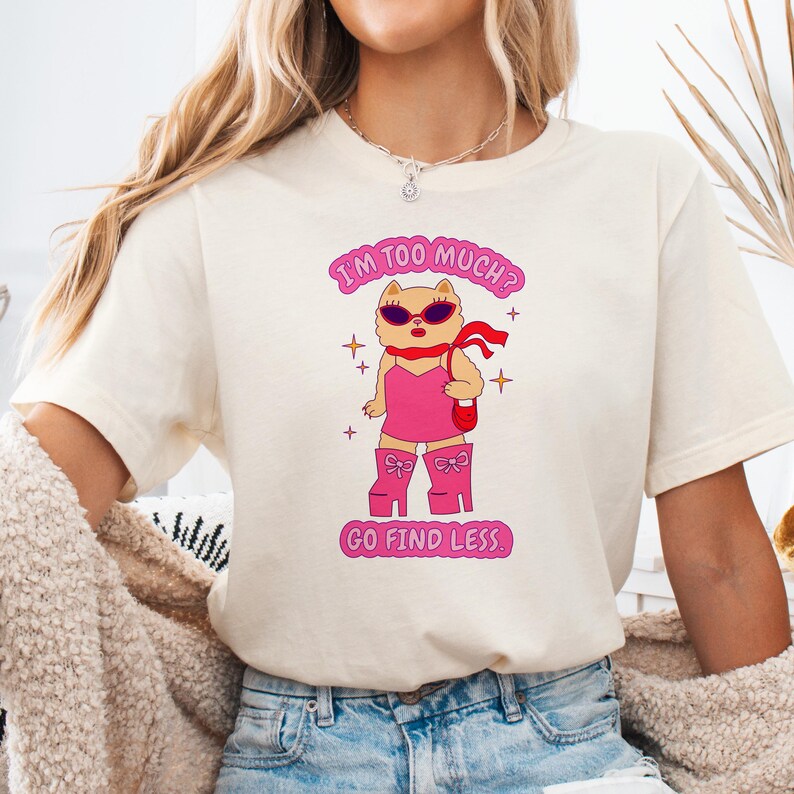 Puede incluir: Una camiseta blanca con un gato rosa de dibujos animados que lleva gafas de sol y botas rosas. El texto de la camiseta dice "I'm too much? Go find less."