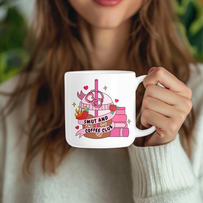 May include: White ceramic mug featuring a colorful illustration of a coffee drink with a pink heart-shaped design, a stack of books, and the text "SMUT AND COFFEE CLUB". The mug has a handle and is held by a person.