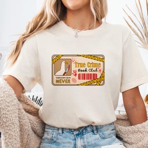 Pu&ograve; includere: Una maglietta bianca con una grafica di un nastro di scena del crimine con il testo "True Crime Book Club" e un'oca bianca che tiene un coltello insanguinato. Il testo "expiration date NEVER" &egrave; sotto la grafica.