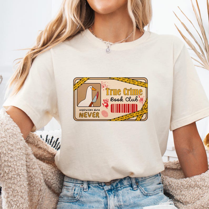 Pu&ograve; includere: Una maglietta bianca con una grafica di un nastro di scena del crimine con il testo "True Crime Book Club" e un'oca bianca che tiene un coltello insanguinato. Il testo "expiration date NEVER" &egrave; sotto la grafica.