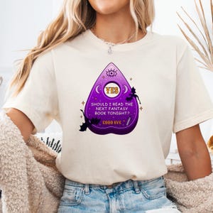 Op de afbeelding: Een wit T-shirt met een paarse en gele afbeelding van een Ouija-bord met de tekst "Should I read the next fantasy book tonight? YES GOOD BYE".
