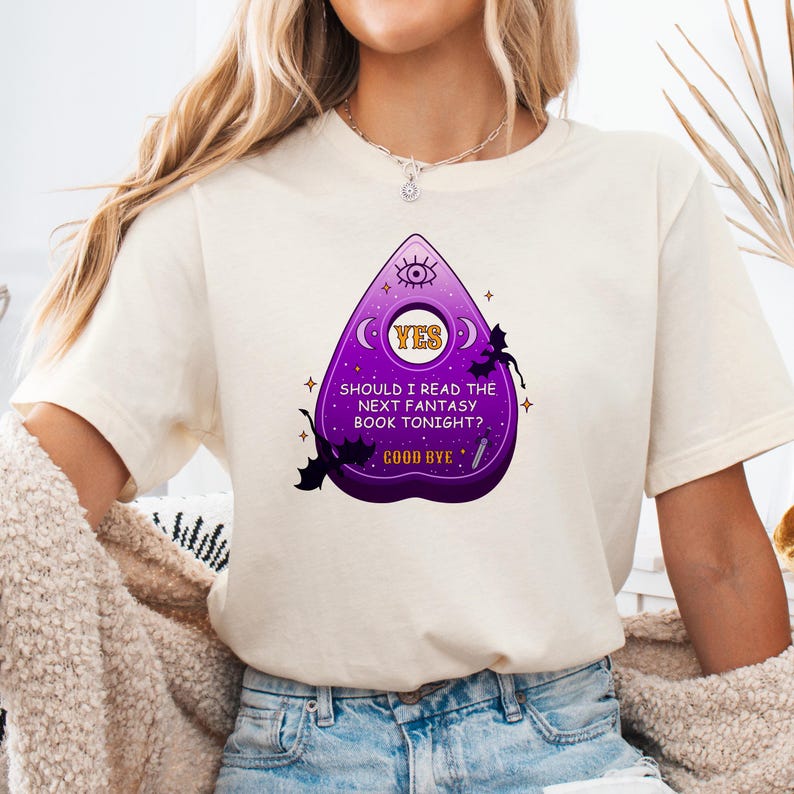 Op de afbeelding: Een wit T-shirt met een paarse en gele afbeelding van een Ouija-bord met de tekst "Should I read the next fantasy book tonight? YES GOOD BYE".