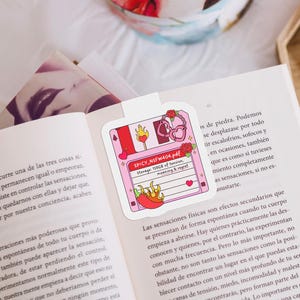 May include: A pink floppy disk-shaped bookmark with a playful design. It features a red chili pepper, hearts, and the text "SPICY_NSFW404.pdf". The bookmark is on an open book.