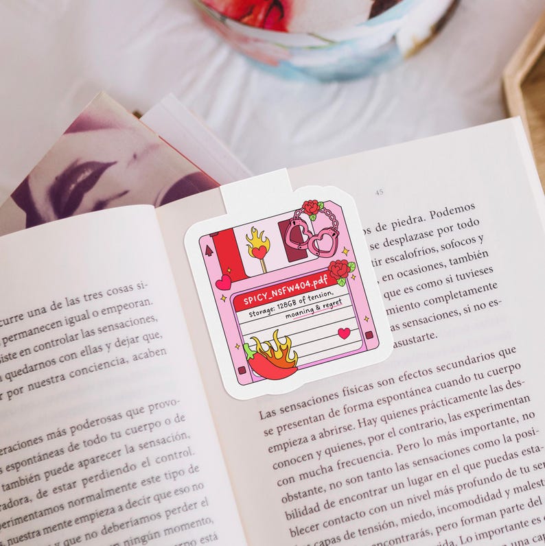 May include: A pink floppy disk-shaped bookmark with a playful design. It features a red chili pepper, hearts, and the text "SPICY_NSFW404.pdf". The bookmark is on an open book.