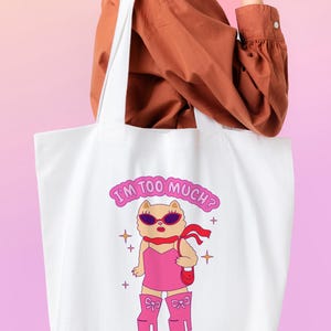 Puede incluir: Bolsa de tela blanca con un gato de dibujos animados rosa que lleva gafas de sol y un vestido rosa. El texto en la bolsa dice "I'm too much? Go find less."