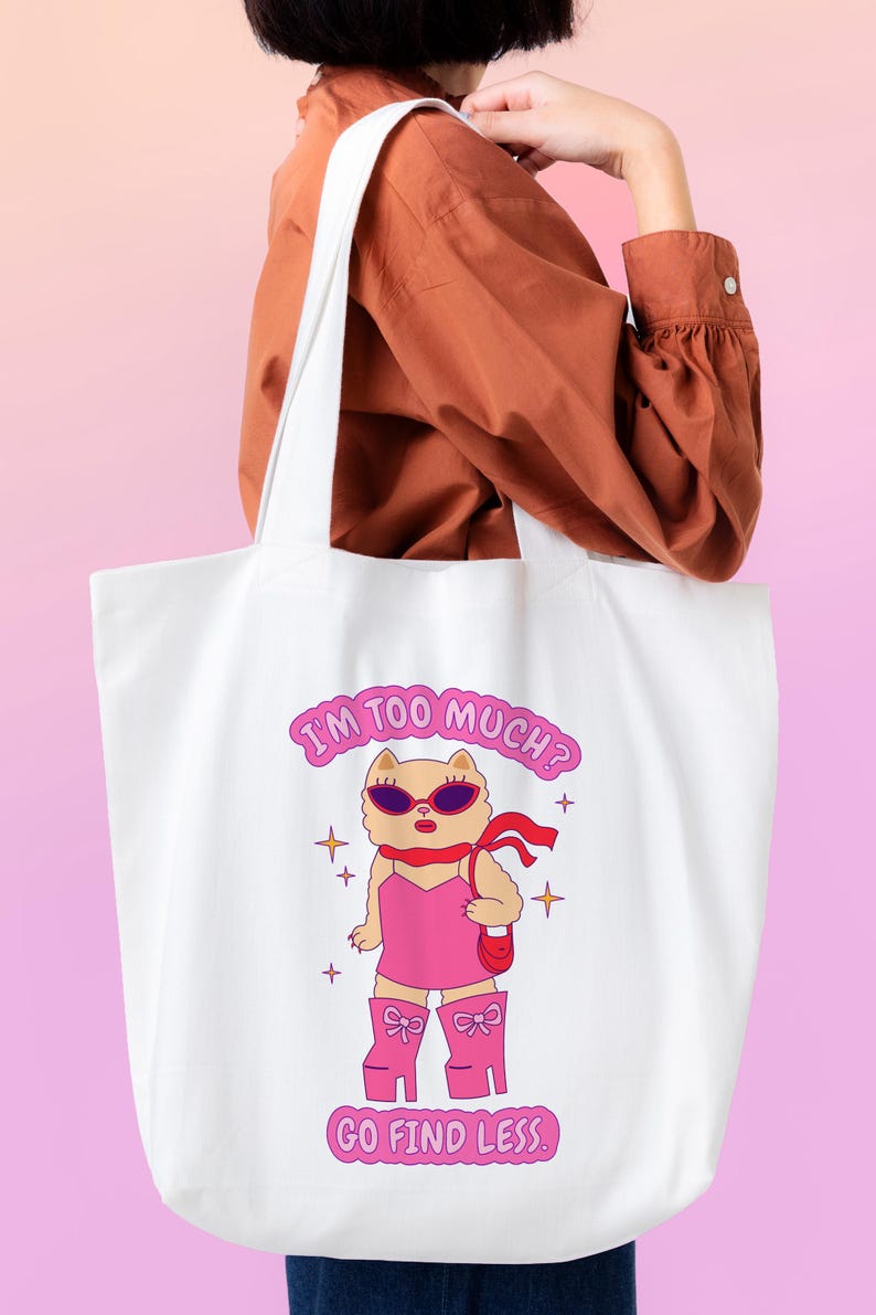 Puede incluir: Bolsa de tela blanca con un gato de dibujos animados rosa que lleva gafas de sol y un vestido rosa. El texto en la bolsa dice "I'm too much? Go find less."