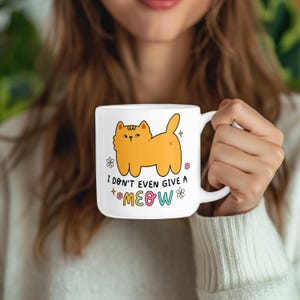以下が含まれることがあります： 白いセラミック製のマグカップに、オレンジ色の漫画の猫と「ニャーも言わない」という文字が書かれています。