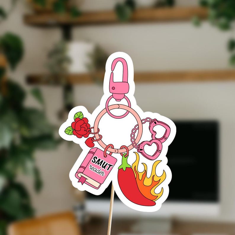 May include: A pink keychain with various charms, including a rose, a book labeled "SMUT reader", a chili pepper with flames, and heart-shaped handcuffs. The keychain has a pink clasp and a ring with a chain.