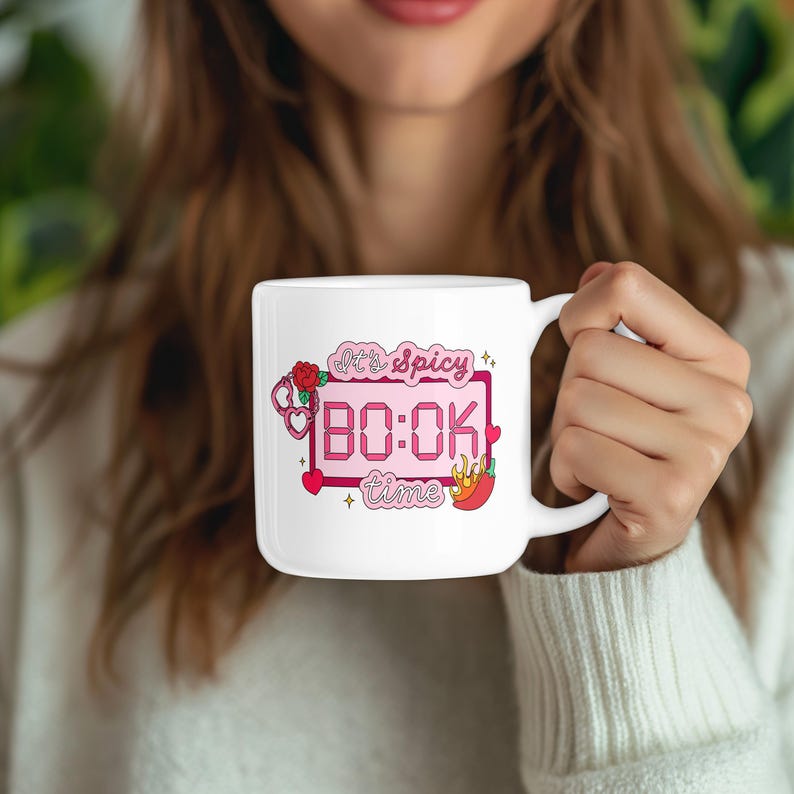 May include: White ceramic mug with a pink graphic that reads "It's Spicy BO:OK time" with hearts, roses, and a chili pepper. The mug is held by a person wearing a white sweater.
