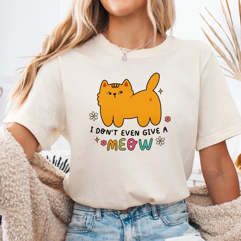 以下が含まれることがあります： オレンジ色の猫の漫画と「I don't even give a meow」というカラフルな文字が書かれた白いTシャツです。