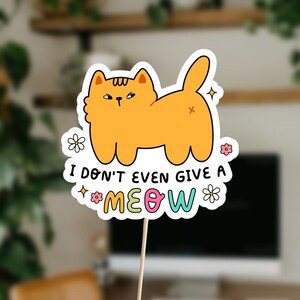 以下が含まれることがあります： 不機嫌そうな表情をしたオレンジ色の漫画の猫が描かれた白いステッカー。猫は視聴者に向かって背中を向けて座っています。猫の下には「ニャーも言わない」というカラフルな文字が書かれています。