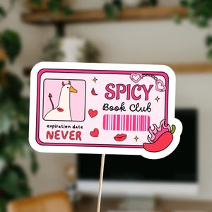 Pu&ograve; includere: Una carta rosa e bianca con un'oca di cartone animato che indossa una fascia con corna di diavolo. La carta dice "Spicy Book Club" con un codice a barre e il testo "expiration date NEVER".