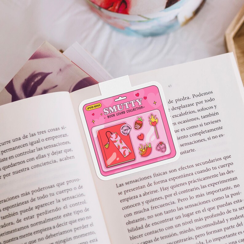 May include: A pink sticker sheet with the text "SMUTTY - BOOK LOVED COLLECTION" and various illustrations, including a book, a strawberry, and a flame. The sticker sheet is placed on an open book.