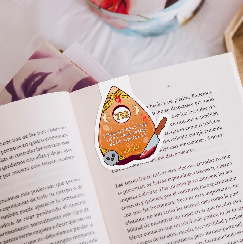 Op de afbeelding: Een gele en oranje Ouija-bord-stijl bladwijzer met een zwarte schedel en een bloedig mes. De bladwijzer zegt "YES" en "Should I read the next true crime book tonight?" met de tekst "Good Bye" eronder.