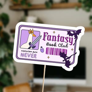 Pu&ograve; includere: Un adesivo viola e bianco con un'oca di cartone animato che indossa una corona e tiene una spada. L'adesivo dice "Fantasy Book Club" e "data di scadenza MAI".