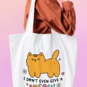 以下が含まれることがあります： 白いトートバッグに、オレンジ色の猫の漫画と「I don't even give a meow」というカラフルな文字が書かれています。
