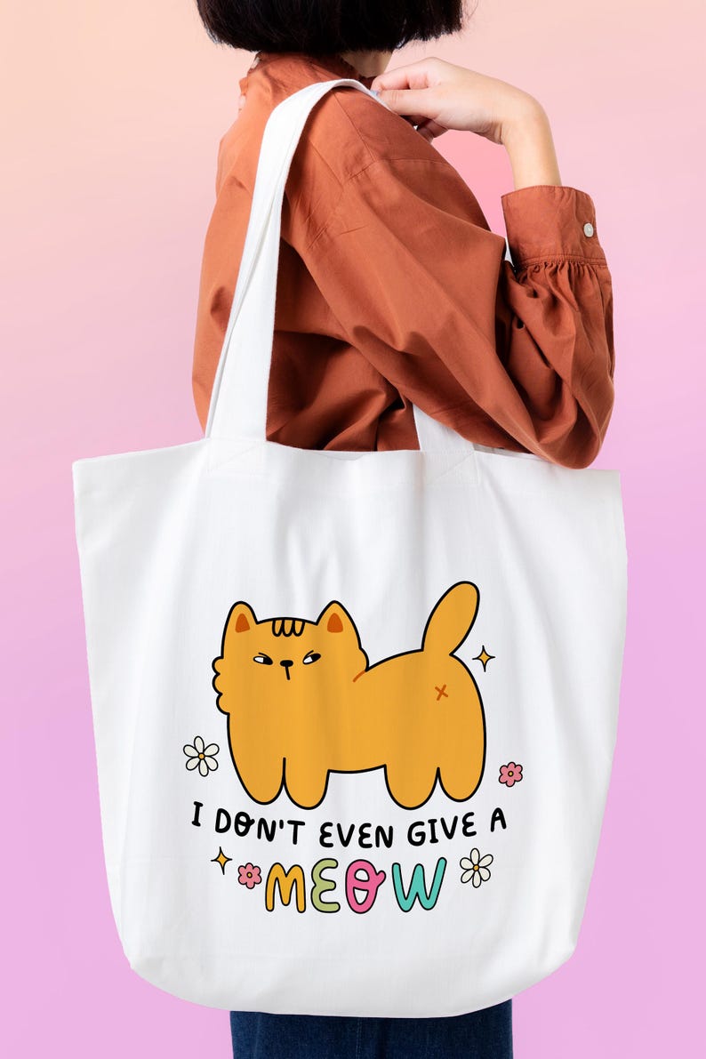 以下が含まれることがあります： 白いトートバッグに、オレンジ色の猫の漫画と「I don't even give a meow」というカラフルな文字が書かれています。