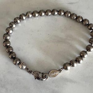 Puede incluir: Una pulsera de cuentas plateadas con cierre de mosquetón. La pulsera presenta pequeñas cuentas redondas ensartadas. El cierre tiene la inscripción "Napier". La pulsera mide aproximadamente 18 cm de largo.