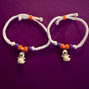 May include: Two white braided cord bracelets featuring a small white ghost charm. Each bracelet is accented with two purple beads and two orange beads, with an additional orange bead positioned at the top of the braided cord loop.