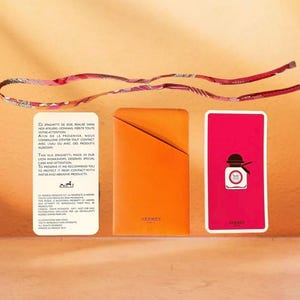 May include: A close-up of a small orange box with the word "HERMES" printed on it. The box is next to a pink card with a black and white illustration of a bottle of perfume. The card also has the word "HERMES" printed on it. The text on the card reads: "C'est une petite gourmandise de la maison Hermes. Afin de le préserver, nous vous conseillons d'éviter tout contact avec l'eau ou avec des produits agressifs."